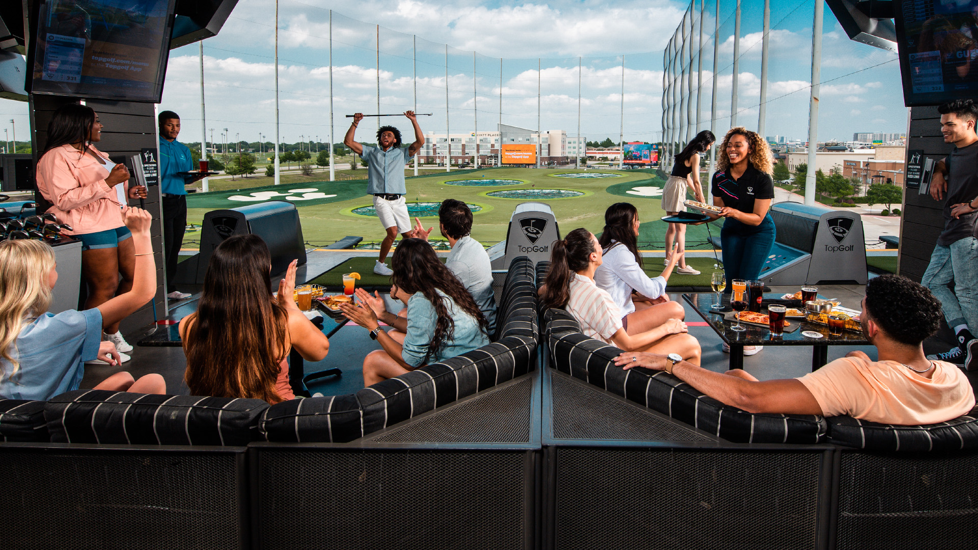 A group seated around a table reacts to a shot at TopGolf in Jacksonville, Florida, with the large green driving range in the background